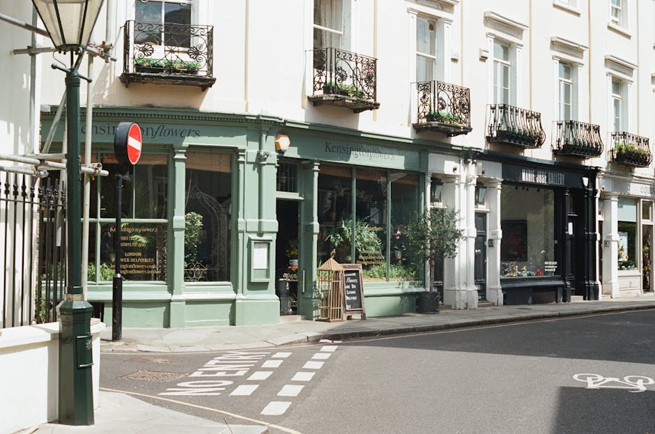A narrow, cobbled residential street in South Kensington lined with terraced houses featuring white and pastel-colored facades, some with small front gardens and potted plants. The street is paved wit