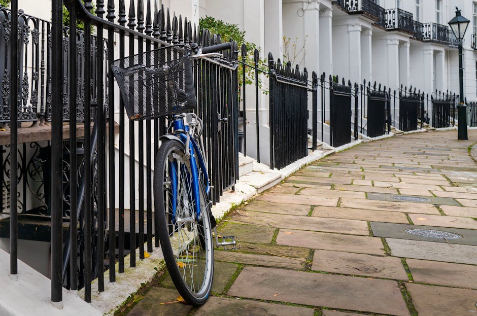 A blue bicycle with a black front basket is parked and locked to a black wrought iron fence along a stone-paved sidewalk outside white terraced residential buildings. The surfaces, including the fence, pavement, and building steps, appear clean and well-maintained, with no visible dirt or debris. The image showcases an orderly street scene in South Kensington, with natural lighting highlighting the shine of the bicycle and the neat appearance of the surroundings. Cleaners South Kensington offers professional surface cleaning and domestic cleaning services to ensure hygiene and tidiness in urban residential areas, as illustrated by the pristine condition of the street scene depicted here.